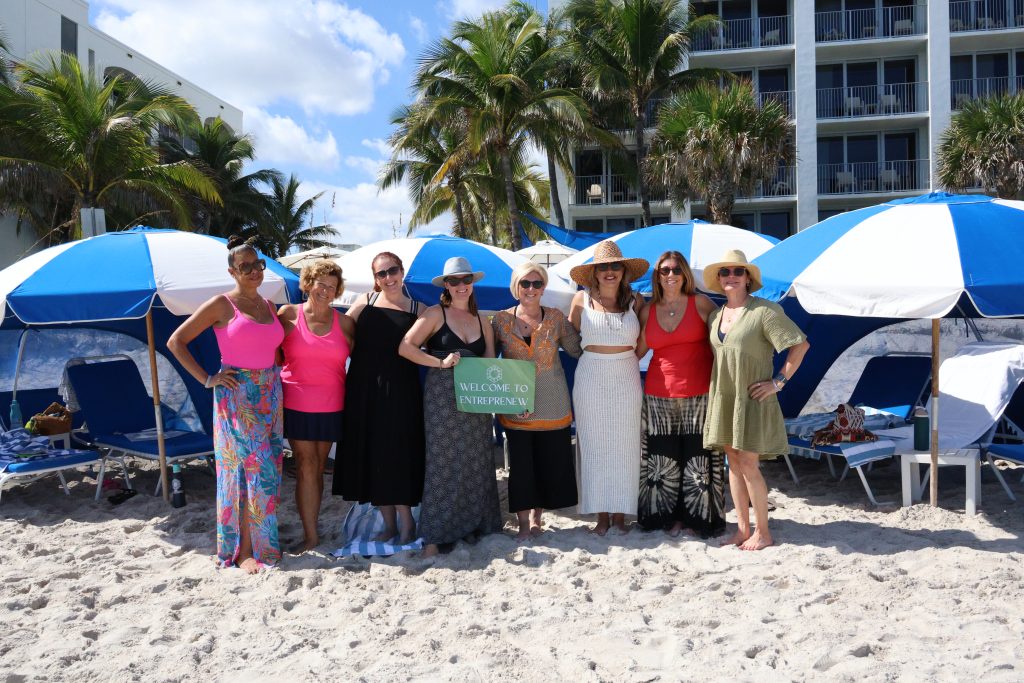 A group of women stand in front of resort beach cabanas holding a 'welcome to entreprenew' sign