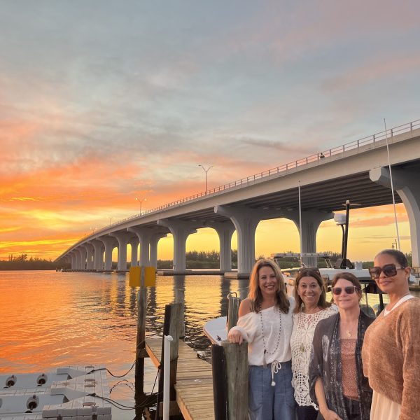 A group of women stand together on a dock with a sunset and arched bridge in the background