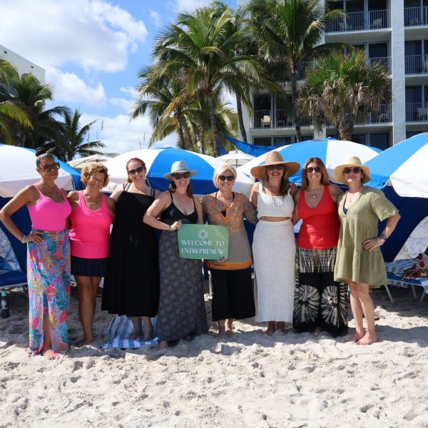 A group of women stand in front of resort beach cabanas holding a 'welcome to entreprenew' sign