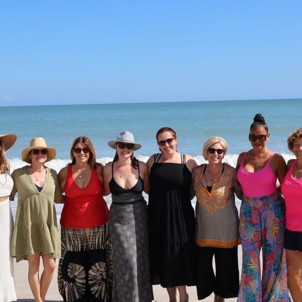 Group of women stand with arms around one another on the beach with the ocean behind them